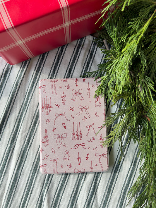 amazon kindle paperwhite case with a design of red bows, candles, and bells on a baby pink background.